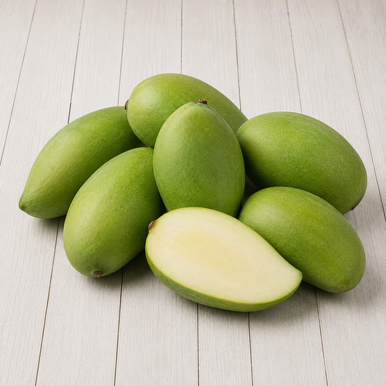 Several green mangoes arranged on a light wood plank surface, with one mango cut in half to show the pale yellow-green flesh.