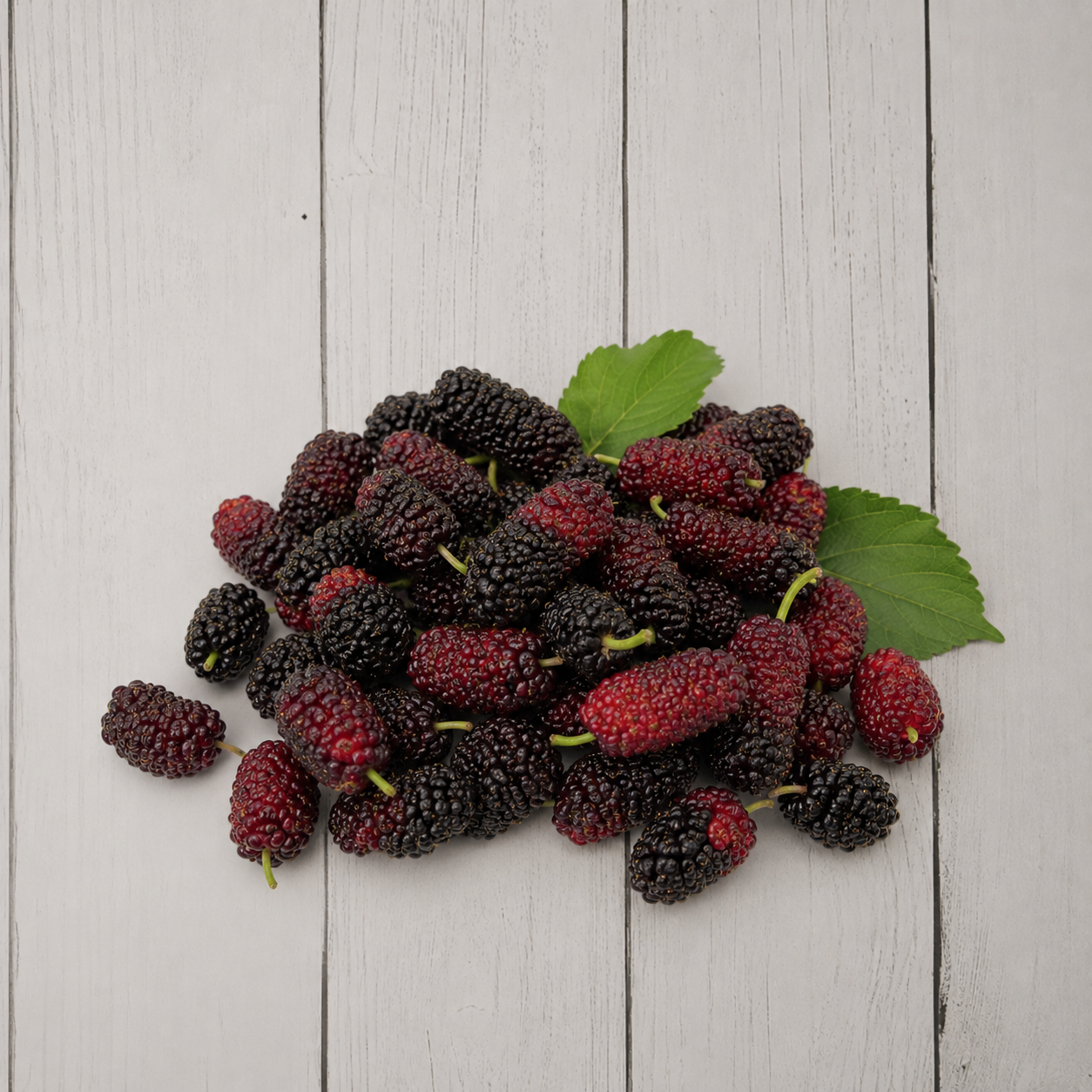 A pile of ripe dark red mulberries with a few green leaves, arranged on a white painted wooden plank surface.