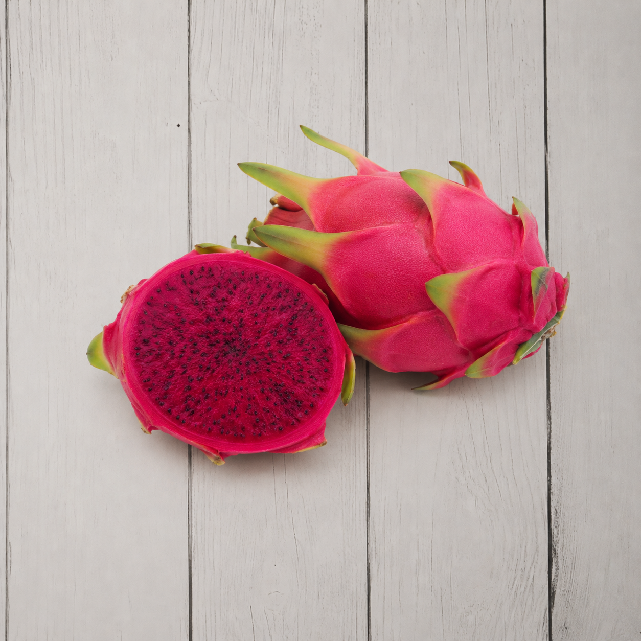 Two dragon fruit fruits on a light gray wooden plank background, with one whole fruit and one sliced open to show the bright magenta flesh and dark seeds.