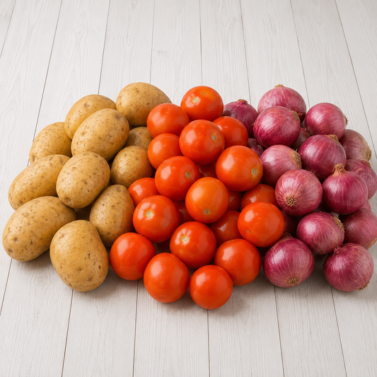 A pile of fresh vegetables on a white wooden surface, including tan potatoes on the left, red cherry tomatoes in the center, and red onions on the right.